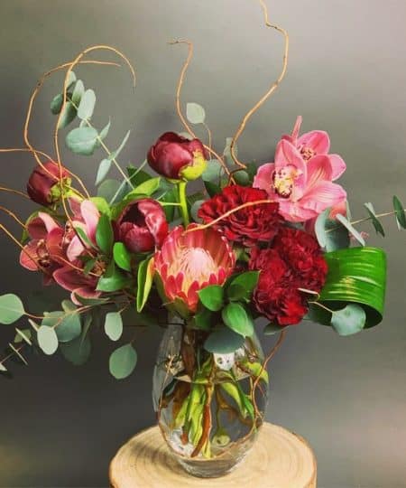 An exotic protea bloom flanked by fluffy red roses and surrounded with blush and wine flowers and eucalyptus. A bouquet suitable for a grand gesture!