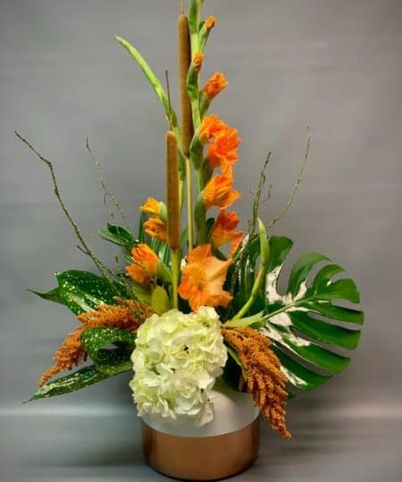 Send a fall gesture with style. Featuring gladiolus and blend of orangey rose gold hues with stunning tropical foliage, this tall and architectural arrangement is sure to impress.
