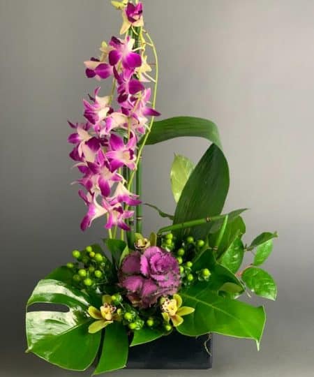A brilliant green container and exotic palm leaf provide the perfect backdrop for purple orchids and a mix of delightful tropical flowers.